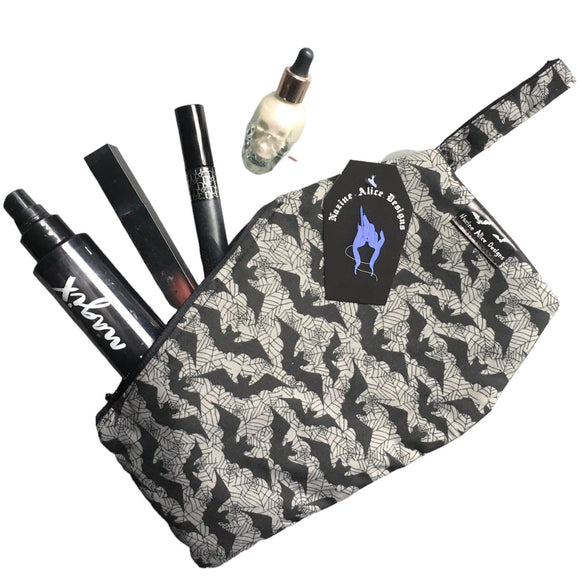 Coffin shaped makeup bag made with gothic fabric. The image shoes various makeup items inside, it can fit a lipstick, mascara, setting spray and highlighter. It has a coffin tag with the Nazine Alice Designs Logo on it.