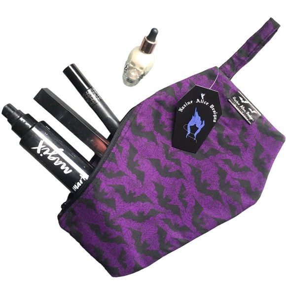 Coffin shaped makeup bag made with gothic fabric. The image shoes various makeup items inside, it can fit a lipstick, mascara, setting spray and highlighter. It has a coffin tag with the Nazine Alice Designs Logo on it.