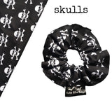 A goth scrunchie in a  skull and crossbones fabric. The fabric has a plain black background with white skulls and crossbones all over as the pattern. This design is called 'Skulls'. The scrunchie has a luxury silk label with the brand name 'Nazine Alice Designs' on it.