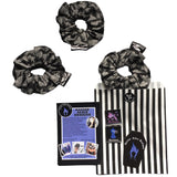 A goth scrunchie in a bats and webs design fabric. The background is a plain grey with spiderwebs and bats flying all over as the pattern. This design is called 'Grey bats and webs'. The scrunchie has a luxury silk label with the brand name 'Nazine Alice Designs' on it. The gift set includes a black and white paper bag, stickers and leaflets.