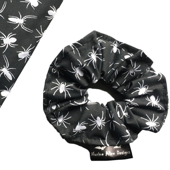 A goth scrunchie in a Spiders fabric, it is a black background with white spiders all over as the pattern. This design is called 'Spiders'. The scrunchie has a luxury silk label with the brand name 'Nazine Alice Designs' on it.