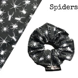 A goth scrunchie in a Spiders fabric, it is a black background with white spiders all over as the pattern. This design is called 'Spiders'. The scrunchie has a luxury silk label with the brand name 'Nazine Alice Designs' on it.