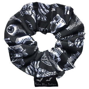 A goth scrunchie in a gothic print fabric, a plain black background with white symbols of skulls, bats, spiders, skeletons, sweets and crows  all over as the pattern. This design is called 'Trick or Treat'. The scrunchie has a luxury silk label with the brand name 'Nazine Alice Designs' on it.