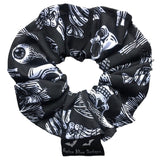 A goth scrunchie in a gothic print fabric, a plain black background with white symbols of skulls, bats, spiders, skeletons, sweets and crows all over as the pattern. This design is called 'Trick or Treat'. The scrunchie has a luxury silk label with the brand name 'Nazine Alice Designs' on it.