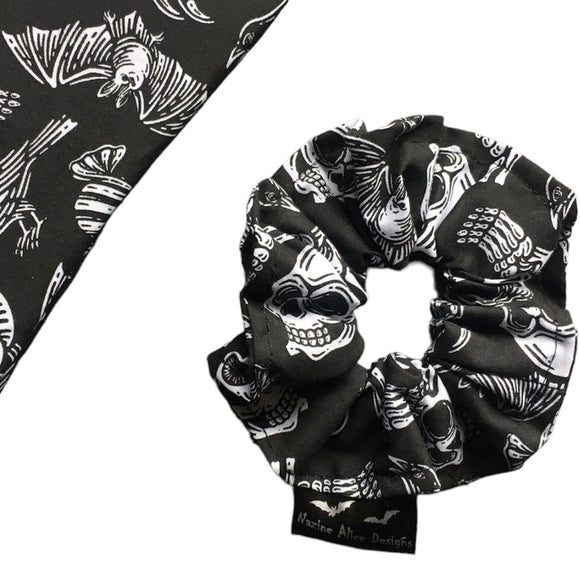 A goth scrunchie in a gothic print fabric, a plain black background with white symbols of skulls, bats, spiders, skeletons, sweets and crows  all over as the pattern. This design is called 'Trick or Treat'. The scrunchie has a luxury silk label with the brand name 'Nazine Alice Designs' on it.