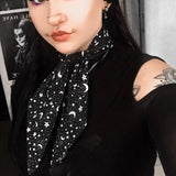 Black and white pentagrams scarf being worn by a woman with tattoos and piercings, styling the scarf in a corporate goth inspired look. She is wearing a black cold shoulder top with mesh sleeves and the scarf lays on top creating a smart tie look.