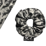 A goth scrunchie in a bats and webs design fabric. The background is a plain grey with spiderwebs and bats flying all over as the pattern. This design is called 'Grey bats and webs'. The scrunchie has a luxury silk label with the brand name 'Nazine Alice Designs' on it.