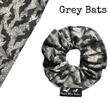 A goth scrunchie in a bats and webs design fabric. The background is a plain grey with spiderwebs and bats flying all over as the pattern. This design is called 'Grey bats and webs'. The scrunchie has a luxury silk label with the brand name 'Nazine Alice Designs' on it.