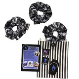 A set of three goth scrunchies in a gothic print fabric, a plain black background with white symbols of skulls, bats, spiders, skeletons, sweets and crows all over as the pattern. This design is called 'Trick or Treat'. The scrunchie has a luxury silk label with the brand name 'Nazine Alice Designs' on it.