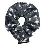 A goth scrunchie in a Spiders fabric, it is a black background with white spiders all over as the pattern. This design is called 'Spiders'. The scrunchie has a luxury silk label with the brand name 'Nazine Alice Designs' on it.