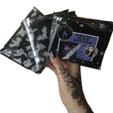 A tattooed hand holding up 3 different scarves folded neatly in recyclable clear packaging bags showing how they'll be received in the post with branded leaflet and stickers included. The patterns include pentagrams, moons, ghosts and bats.