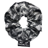 A goth scrunchie in a bats and webs design fabric. The background is a plain grey with spiderwebs and bats flying all over as the pattern. This design is called 'Grey bats and webs'. The scrunchie has a luxury silk label with the brand name 'Nazine Alice Designs' on it.