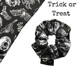 A goth scrunchie in a gothic print fabric, a plain black background with white symbols of skulls, bats, spiders, skeletons, sweets and crows all over as the pattern. This design is called 'Trick or Treat'. The scrunchie has a luxury silk label with the brand name 'Nazine Alice Designs' on it.