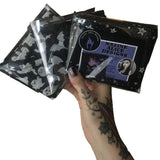 A tattooed hand holding up 3 different scarves folded neatly in recyclable clear packaging bags showing how they&#39;ll be received in the post with branded leaflet and stickers included. The patterns include pentagrams, moons, ghosts and bats.